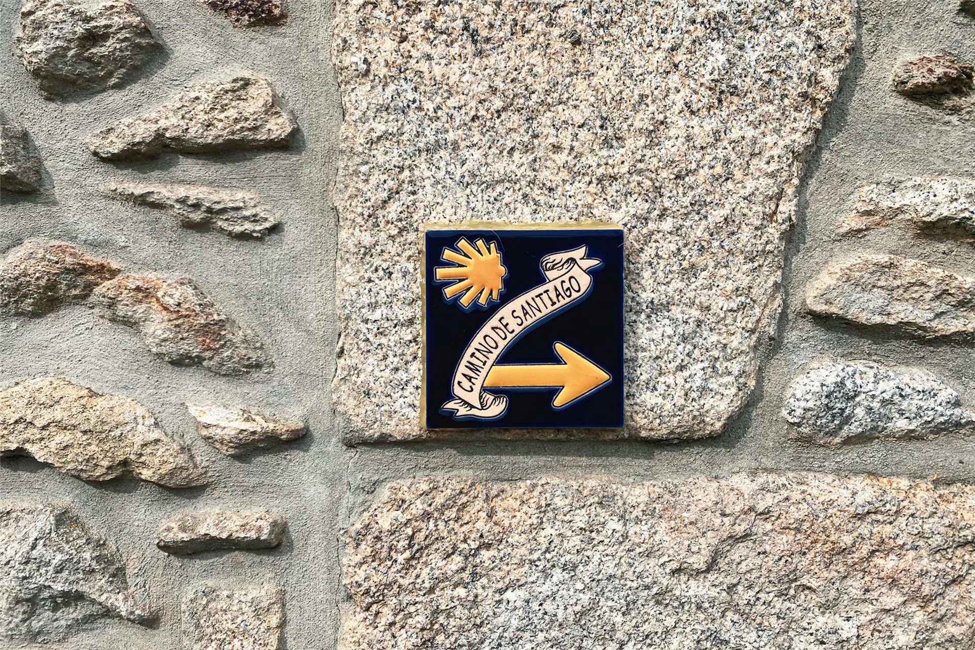 auf einer Steinmauer ist eine quadratische kleine Fließe angebracht auf der eine gelbe Muschel, ein gelber Pfeil und ein Schriftzug "Camino de Santiago" abgebildet sind